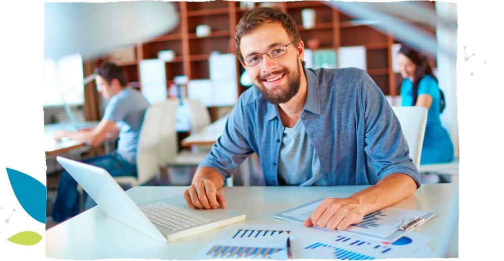Smiling, bearded young man wearing glasses is working on his computer, with papers lying on his white desk.